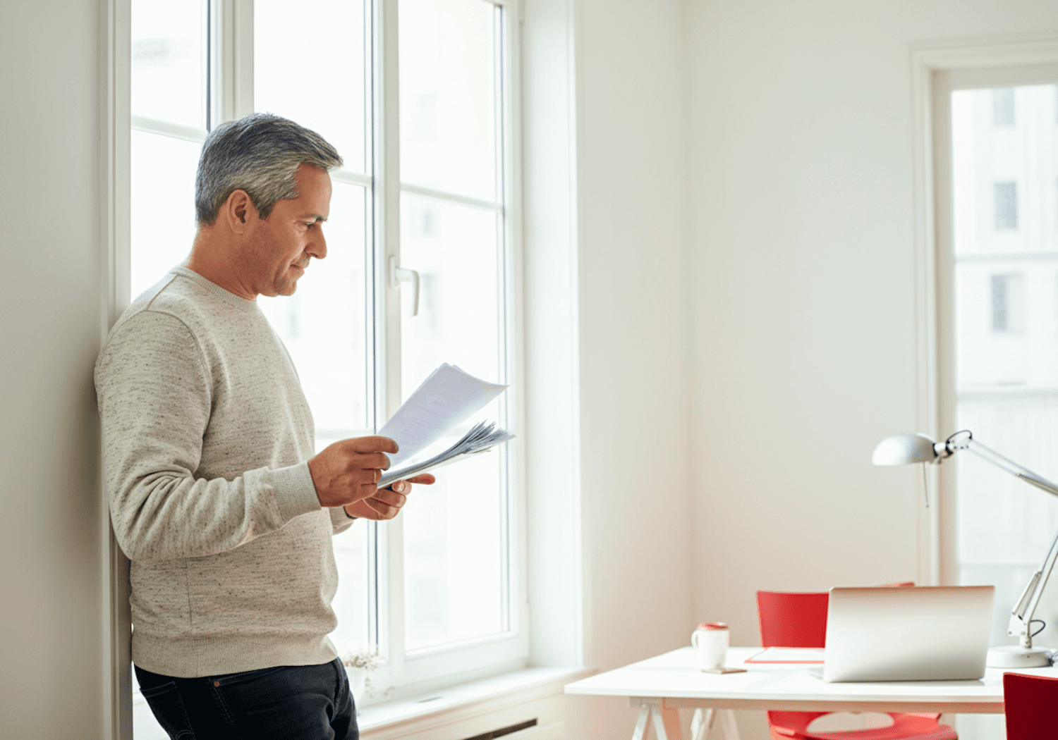 A man reading through printed documents by a bright window
